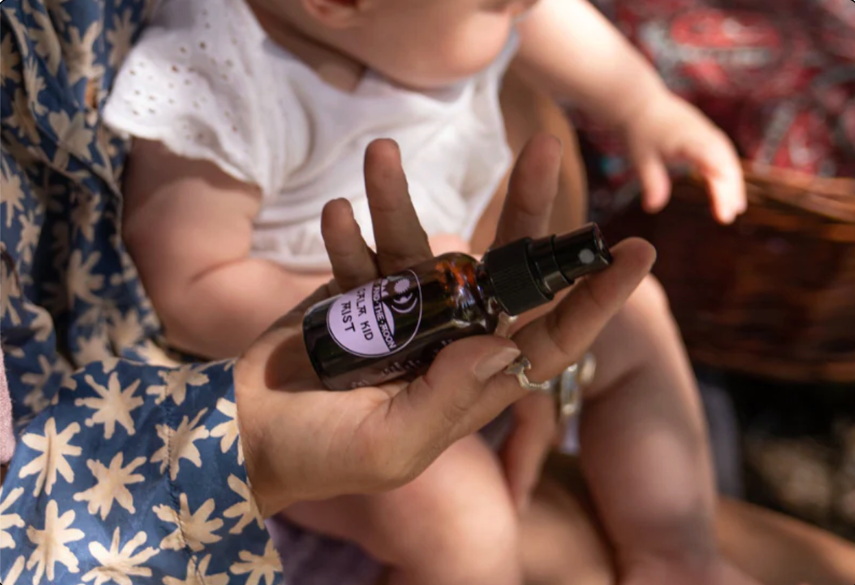 A close-up lifestyle shot of a baby held on a person's lap while the adult's hand holds a small amber spray bottle with a black sprayer. The bottle has a purple and white label with the brand name 'Baba & Yo' and 'Baby Skin Care'.